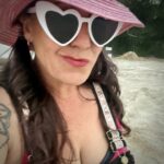 A selfie of the author, smirking into the camera, wearing a pink hat and white rimmed sunglasses that are heart shaped